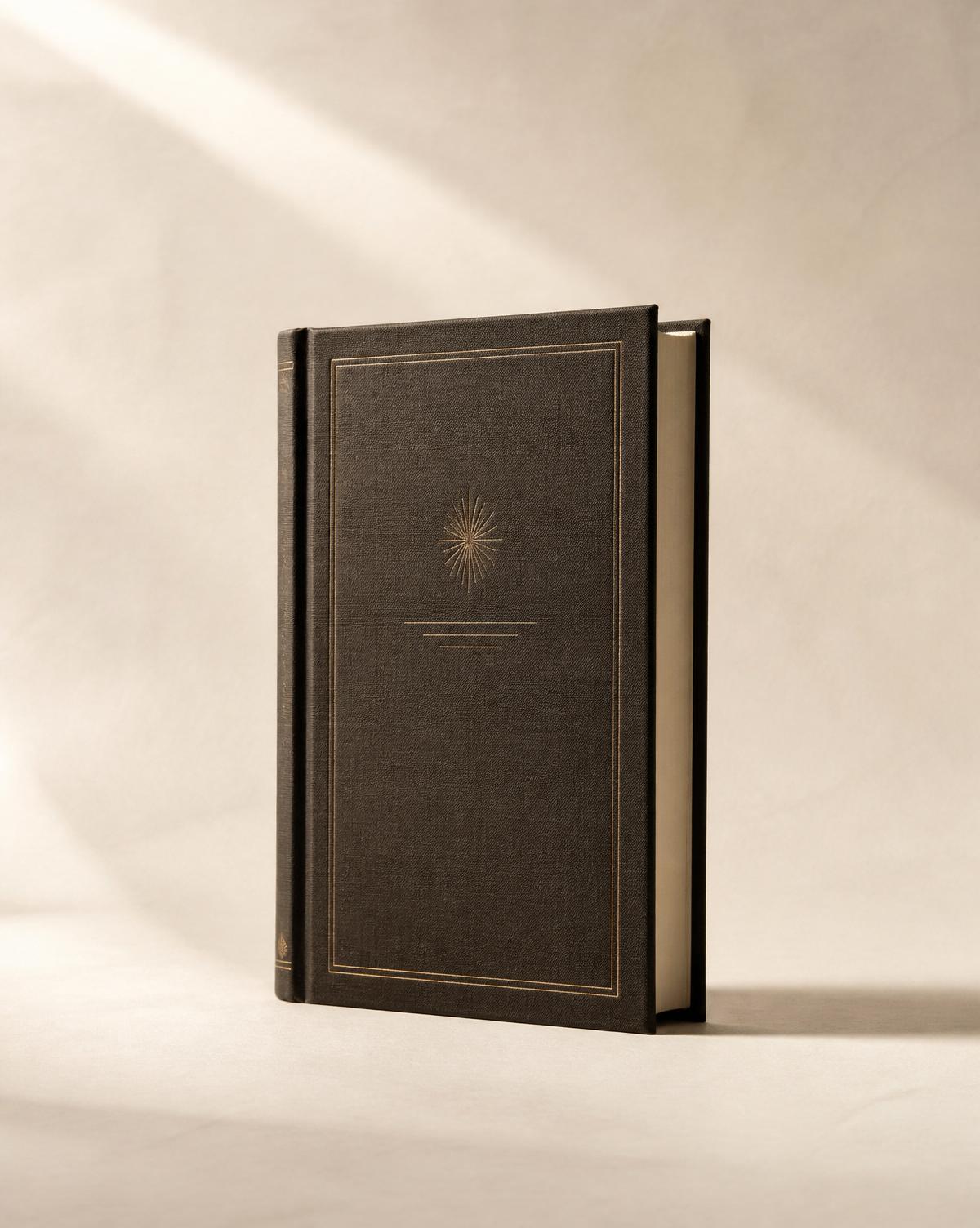 A charcoal linen memoir hardcover with a gold-foil starburst emblem, lit by warm sunlight on ivory paper